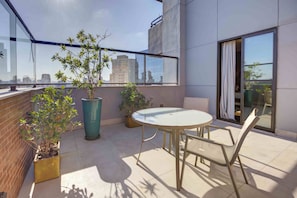 Uma varanda com mesa e cadeiras de vidro, plantas em vasos e vista para o horizonte da cidade.