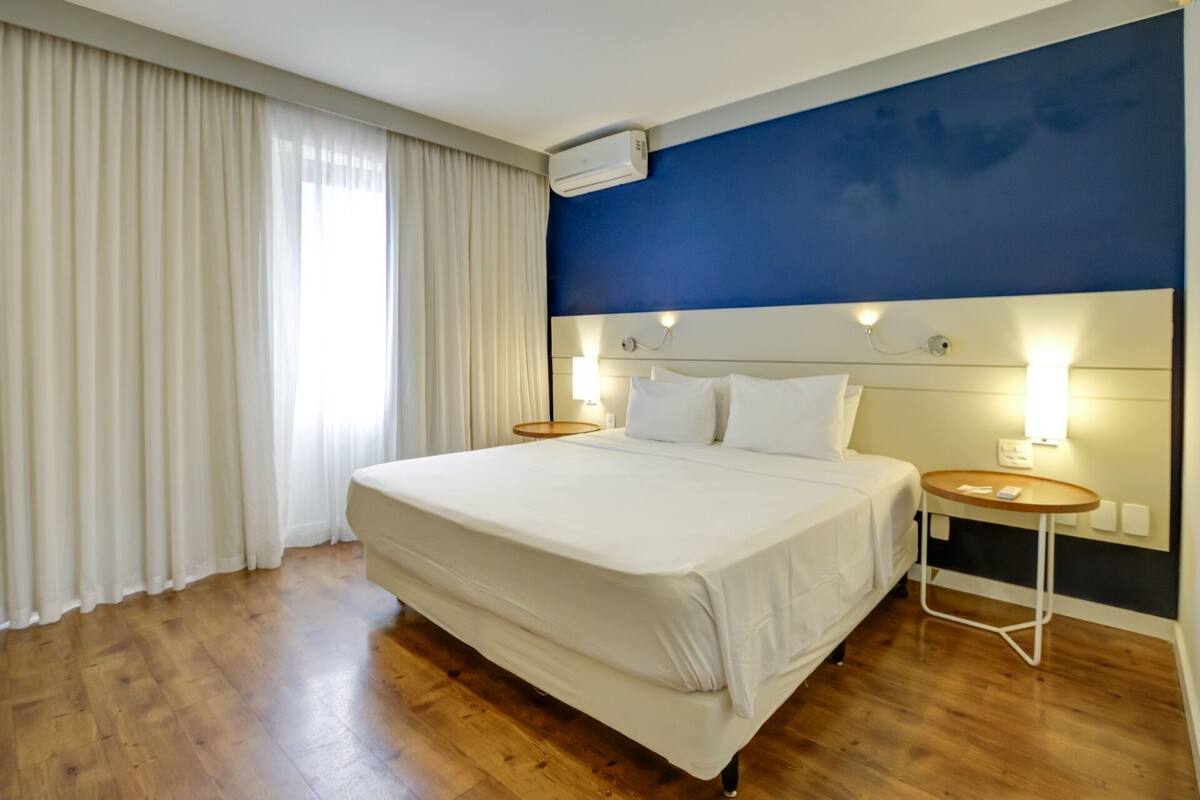 A hotel room with a large bed, a nightstand, a wall-mounted air conditioner, and a window with curtains.
