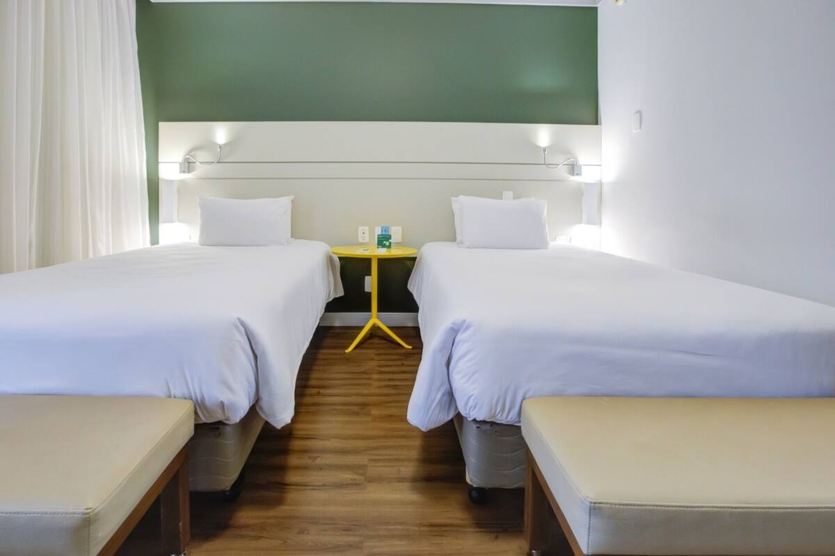 A hotel room with two single beds, a small table with a chair, and a green wall.