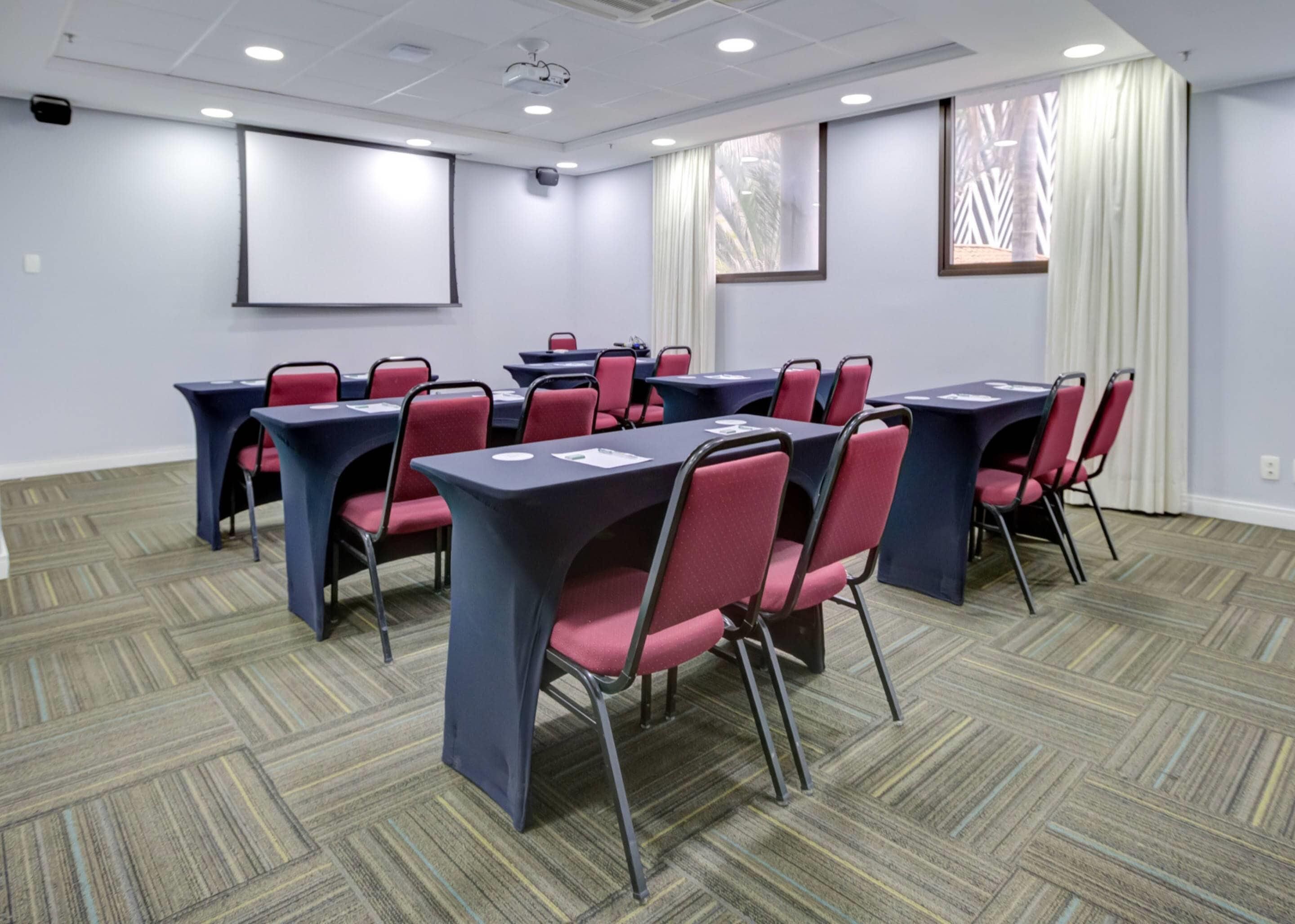 A conference room with tables and chairs, a projector screen, and windows with curtains.