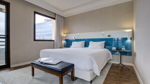 Ein Hotelzimmer mit einem großen Bett, einem Nachttisch, einem kleinen Tisch, einer Bank und Blick auf die Stadt durch ein Fenster.