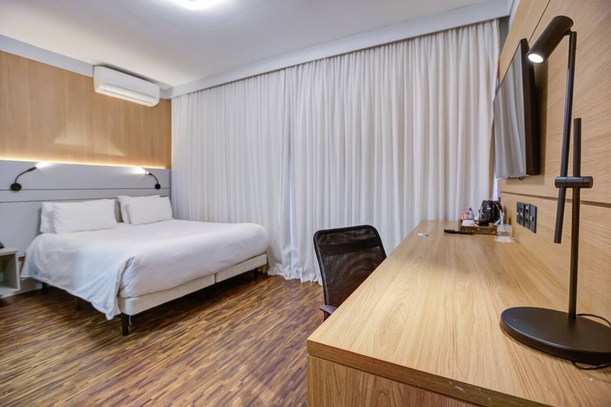 A hotel room with a bed, a desk, a chair, a TV, and a window with curtains.