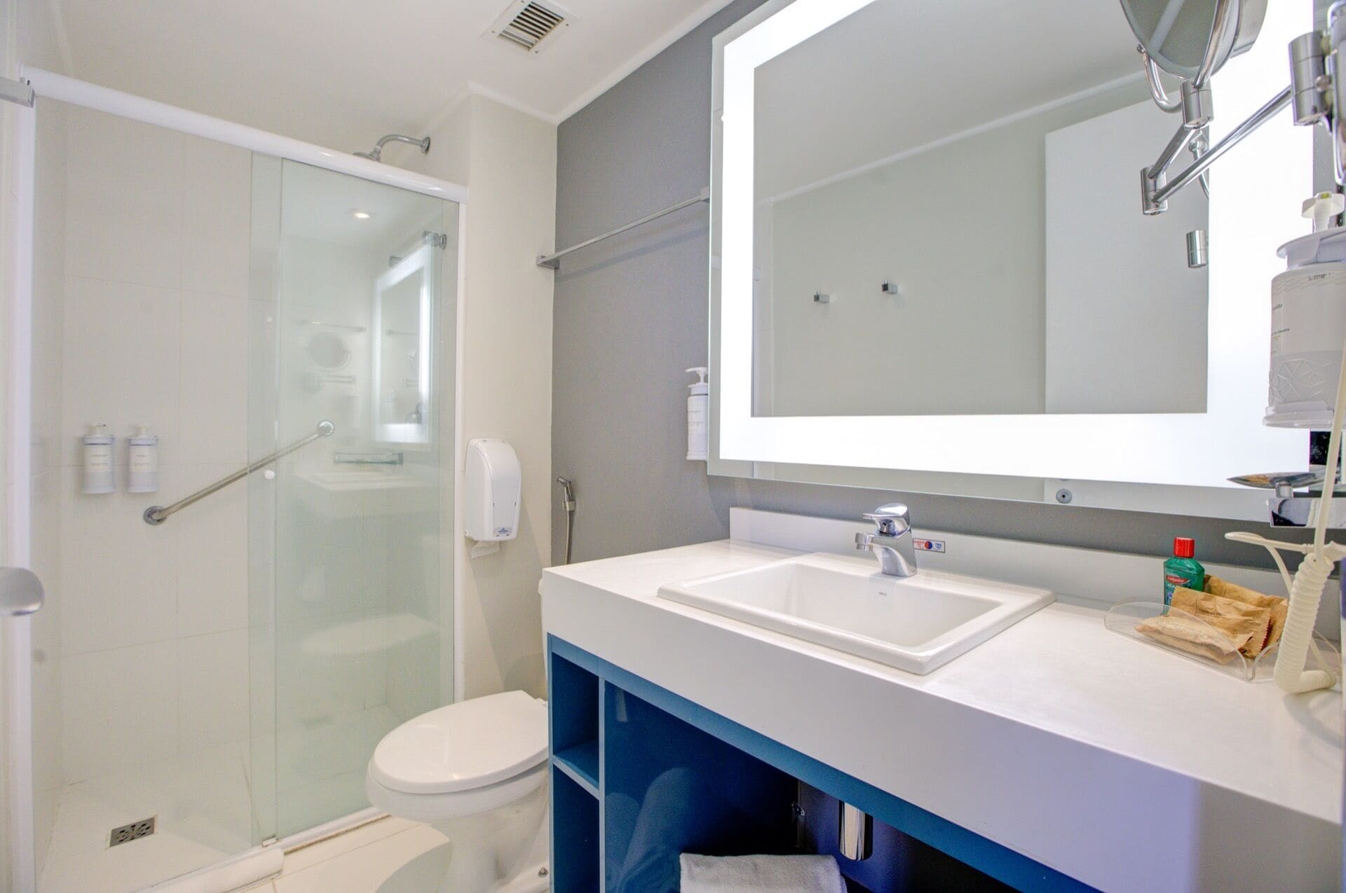 A modern bathroom with a glass-enclosed shower, a white toilet, a large mirror, and a white sink with a blue cabinet.