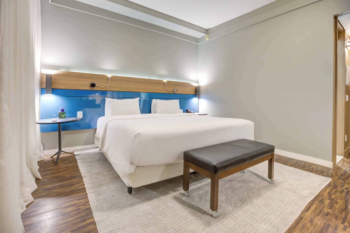 A hotel room with a bed, bedside table, bench, and a headboard with built-in lighting.