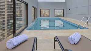 Ein Pool auf dem Dach mit zwei Liegestühlen und Handtüchern, Stadtansichten als Gemälde und Blick auf ein Hochhaus.