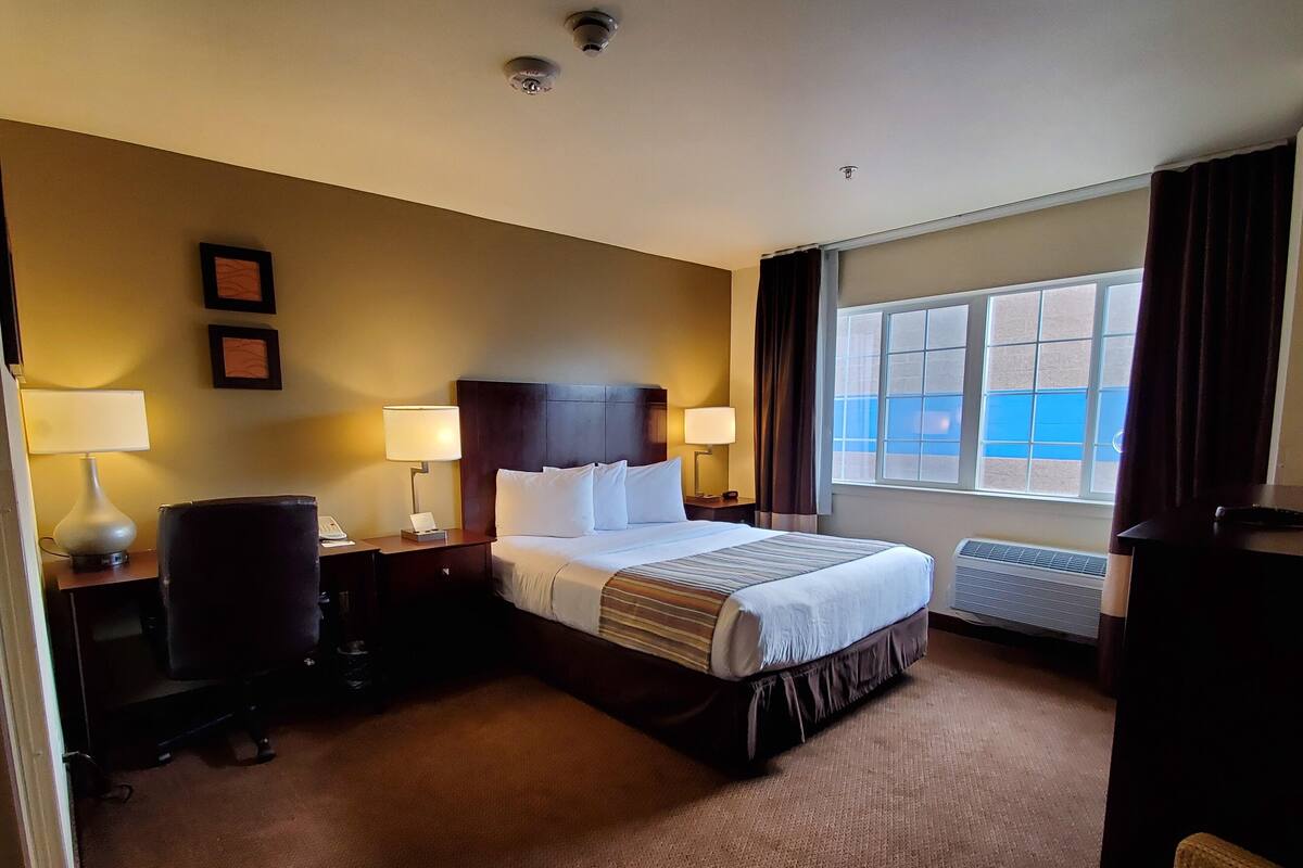 Habitación de hotel con una cama grande, dos lámparas de noche, un escritorio con una silla, una ventana con cortinas y un aire acondicionado instalado en la pared.