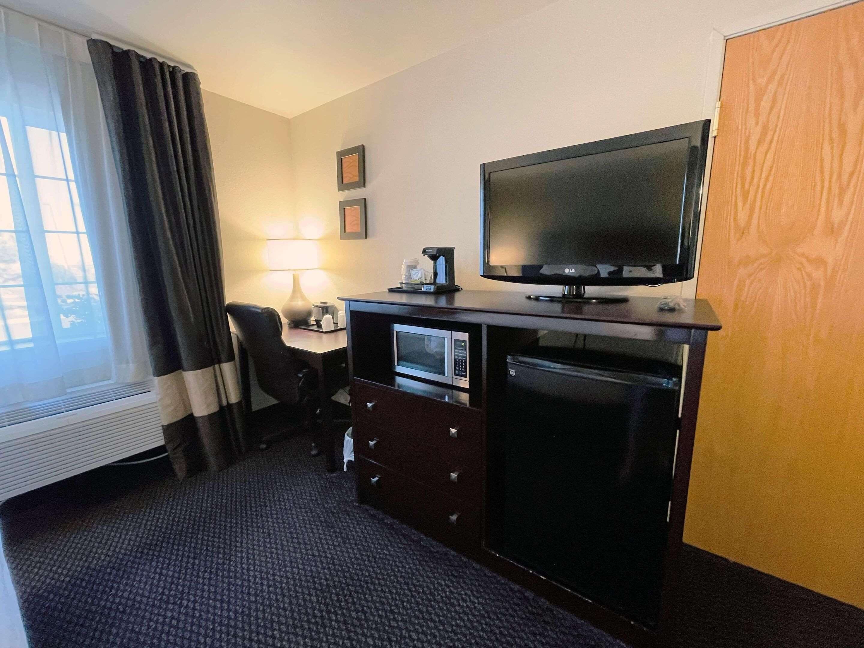 Une chambre d’hôtel équipée d’un téléviseur à écran plat, d’un four à micro-ondes, d’une machine à café, d’un bureau, d’une chaise et d’une fenêtre avec des rideaux.