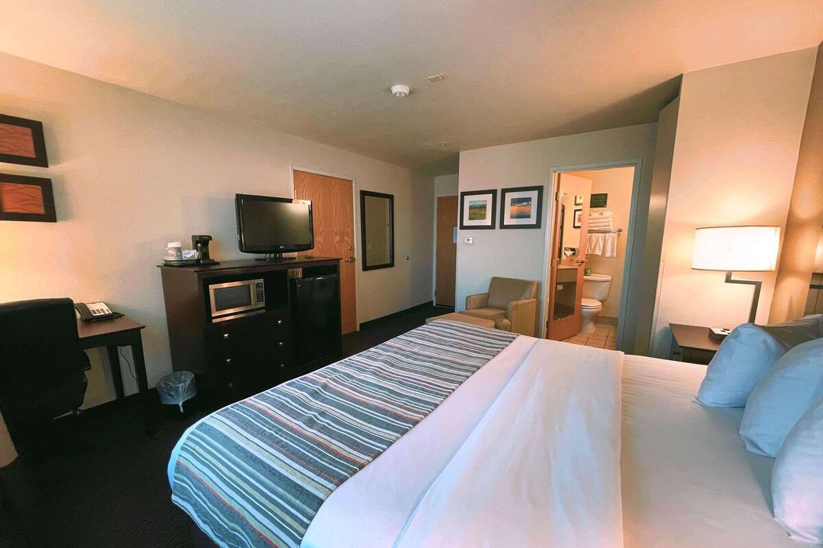 Habitación de hotel con cama, escritorio con teléfono, televisión, microondas, silla y baño con inodoro y espejo.