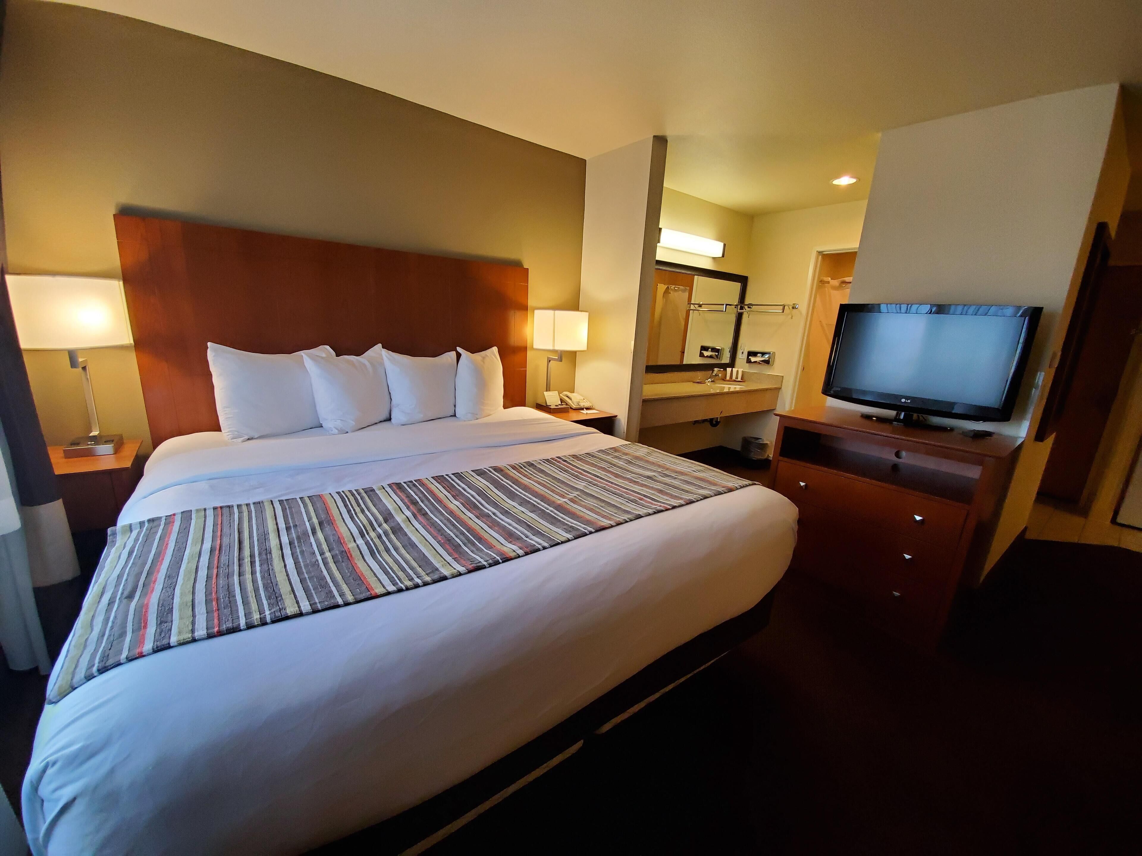 A hotel room with a large bed, a television, a bedside table with a lamp, a mirror, and a bathroom with a sink and mirror.