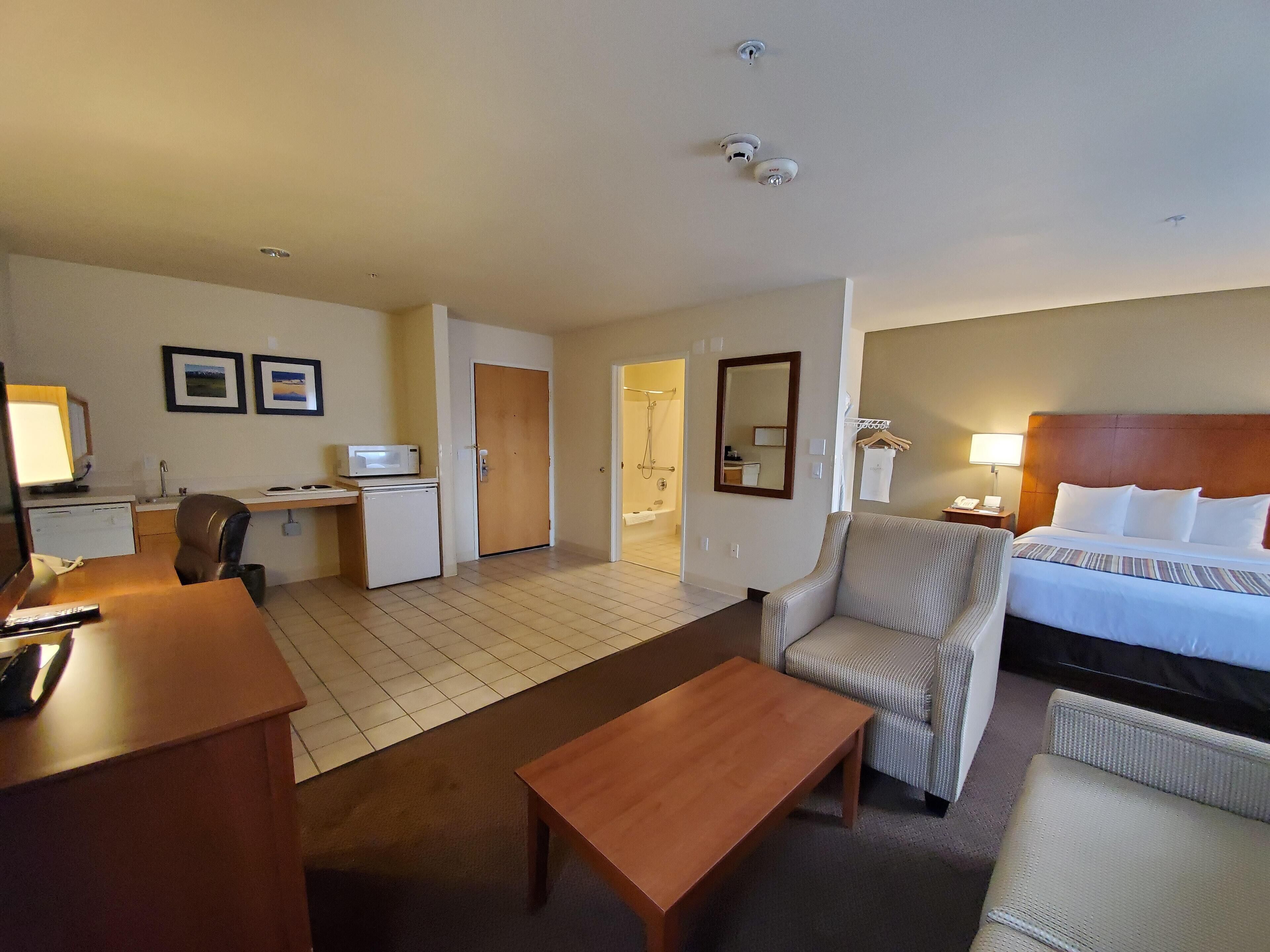 A hotel room with a bed, a desk with a computer, a chair, a small table, and a bathroom visible through an open door.