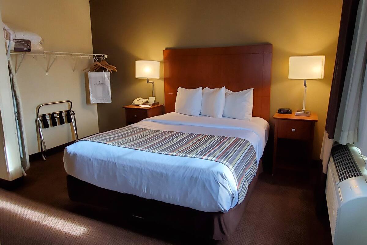 Habitación de hotel con una cama grande, dos mesitas de noche con lámparas, un teléfono, un perchero con un bolso y un equipo de aire acondicionado.