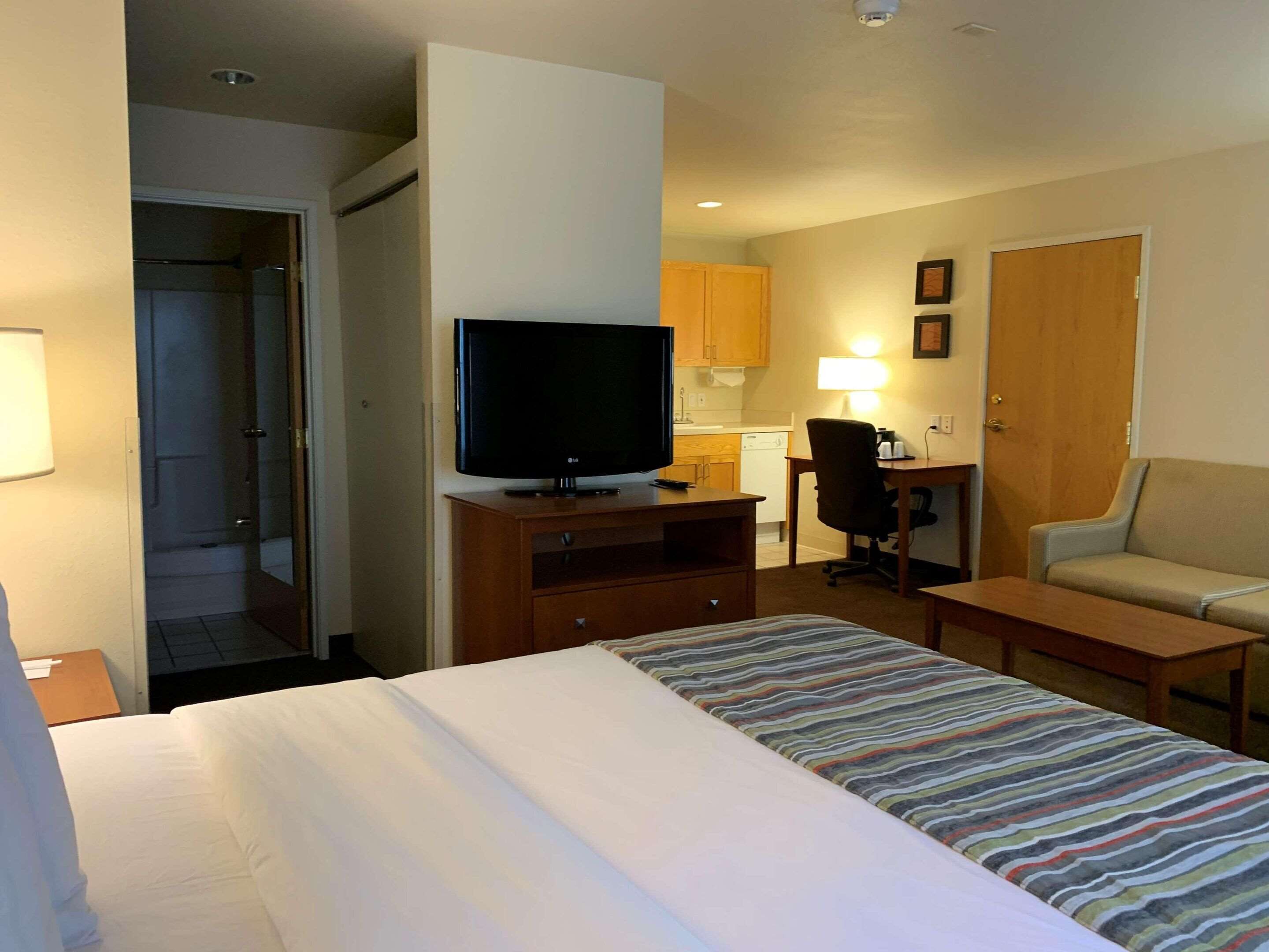 A hotel room with a bed, a TV on a wooden stand, a desk with a chair, a sofa, and a kitchenette in the background.