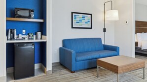 Habitación de hotel pequeña con un sofá azul, una mesita, microondas, refrigerador y un cuadro en la pared.