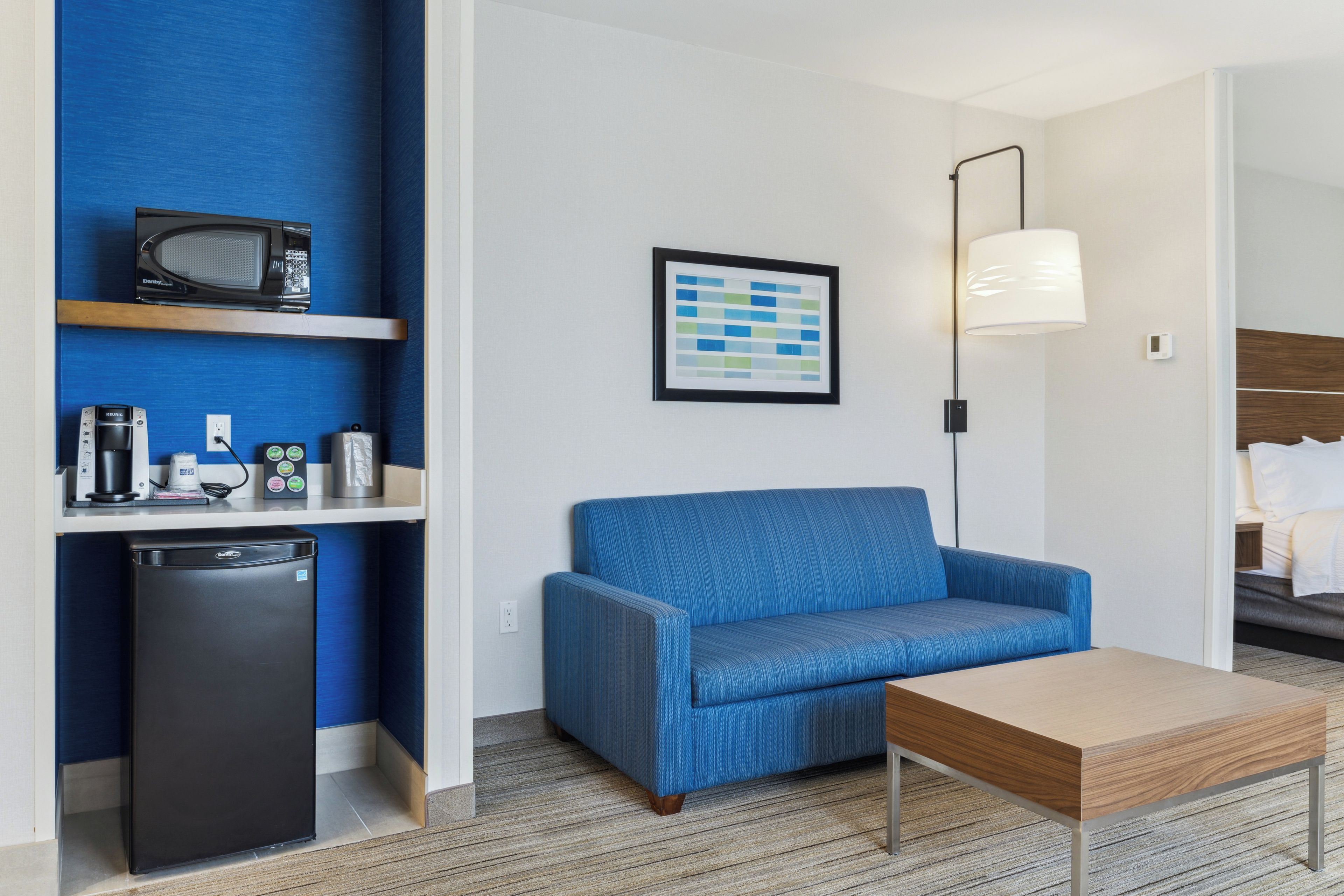 Kompakti hotellihuone, jossa on sininen sohva, pieni pöytä, mikroaaltouuni, jääkaappi ja seinällä oleva kehystetty kuva.