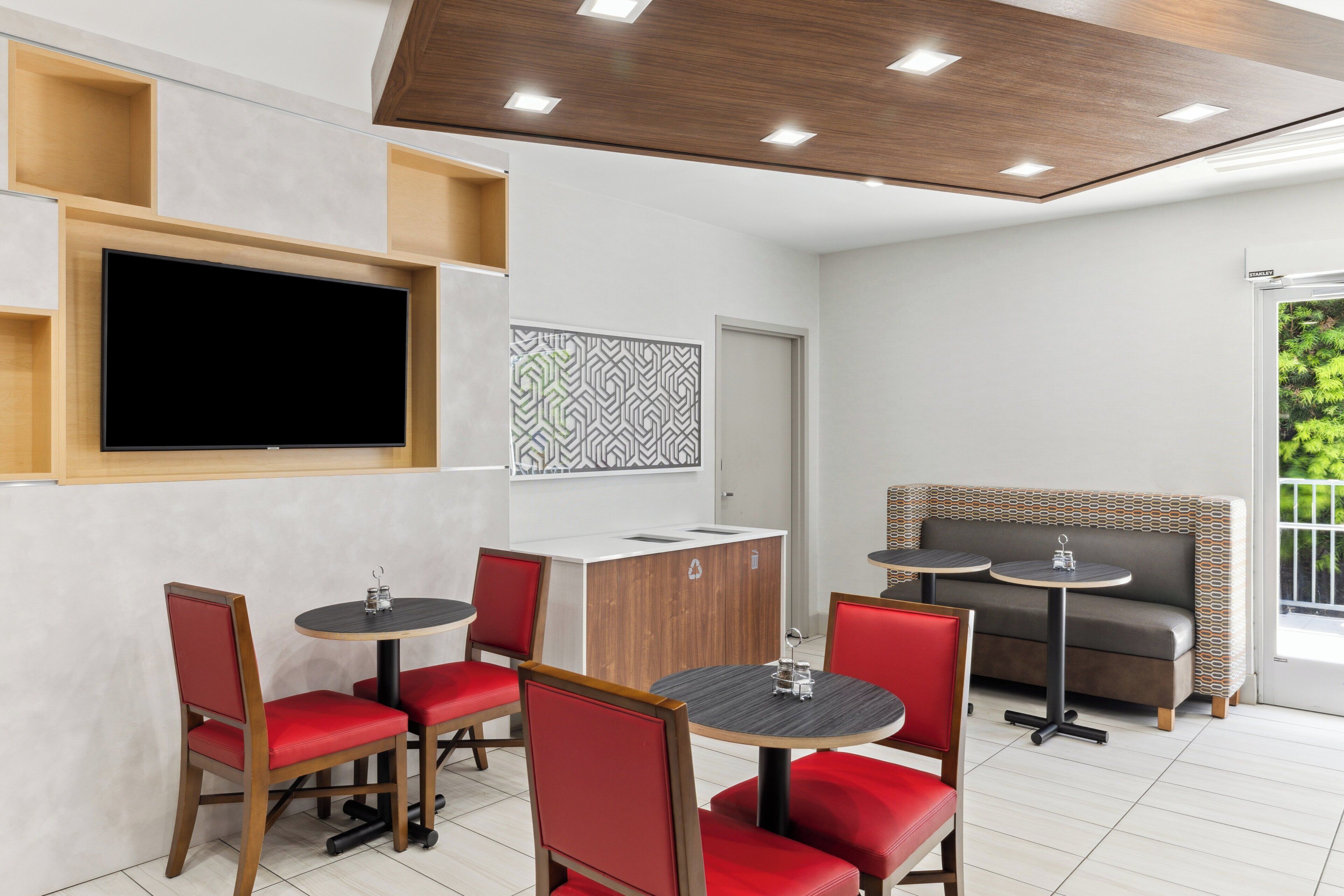Un comedor moderno con sillas rojas, mesas redondas, un aparador de madera y un televisor de pantalla plana montado en la pared.
