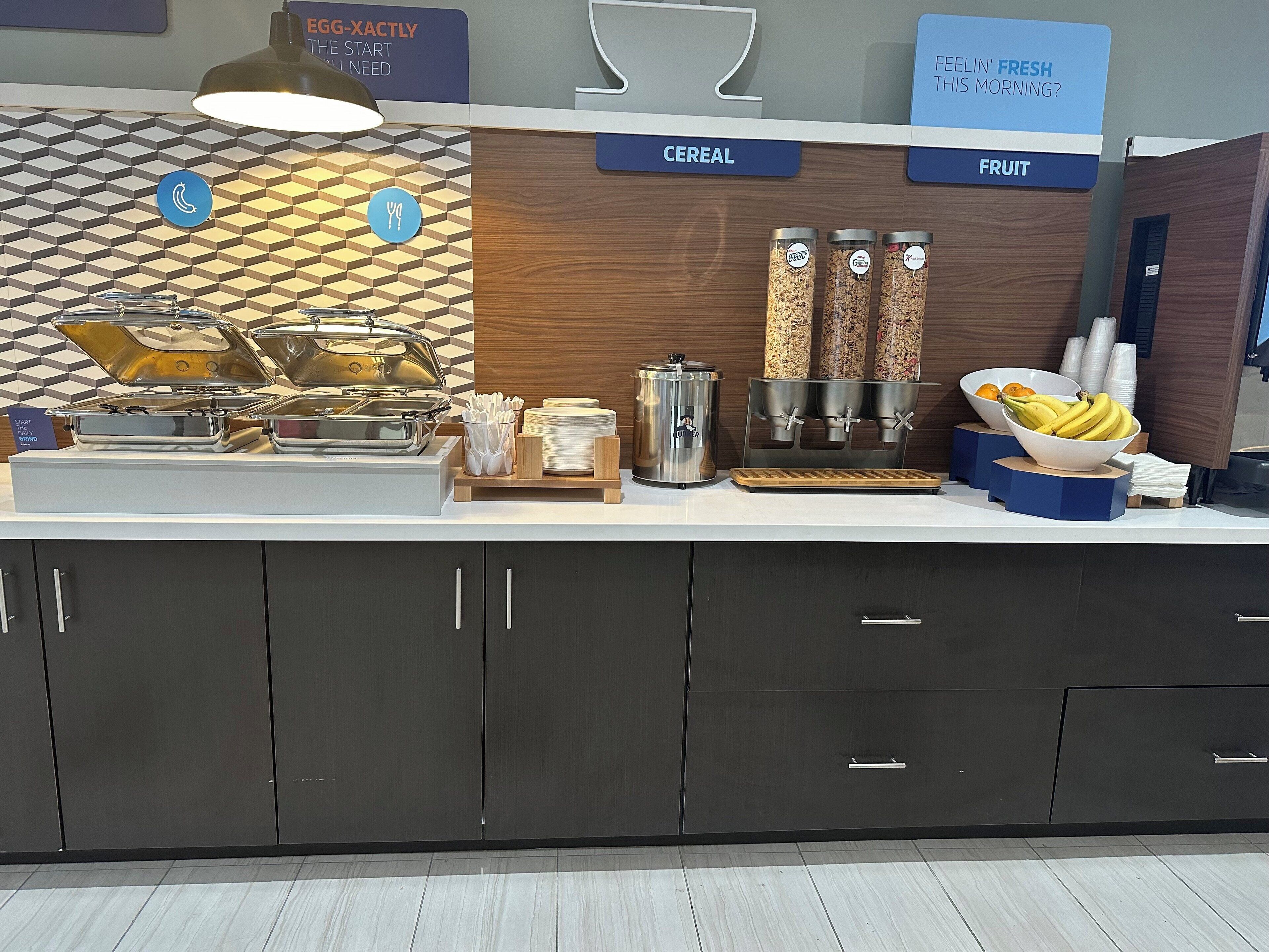 A breakfast buffet area with labeled sections for cereal and fruit, including a coffee machine and various food containers.