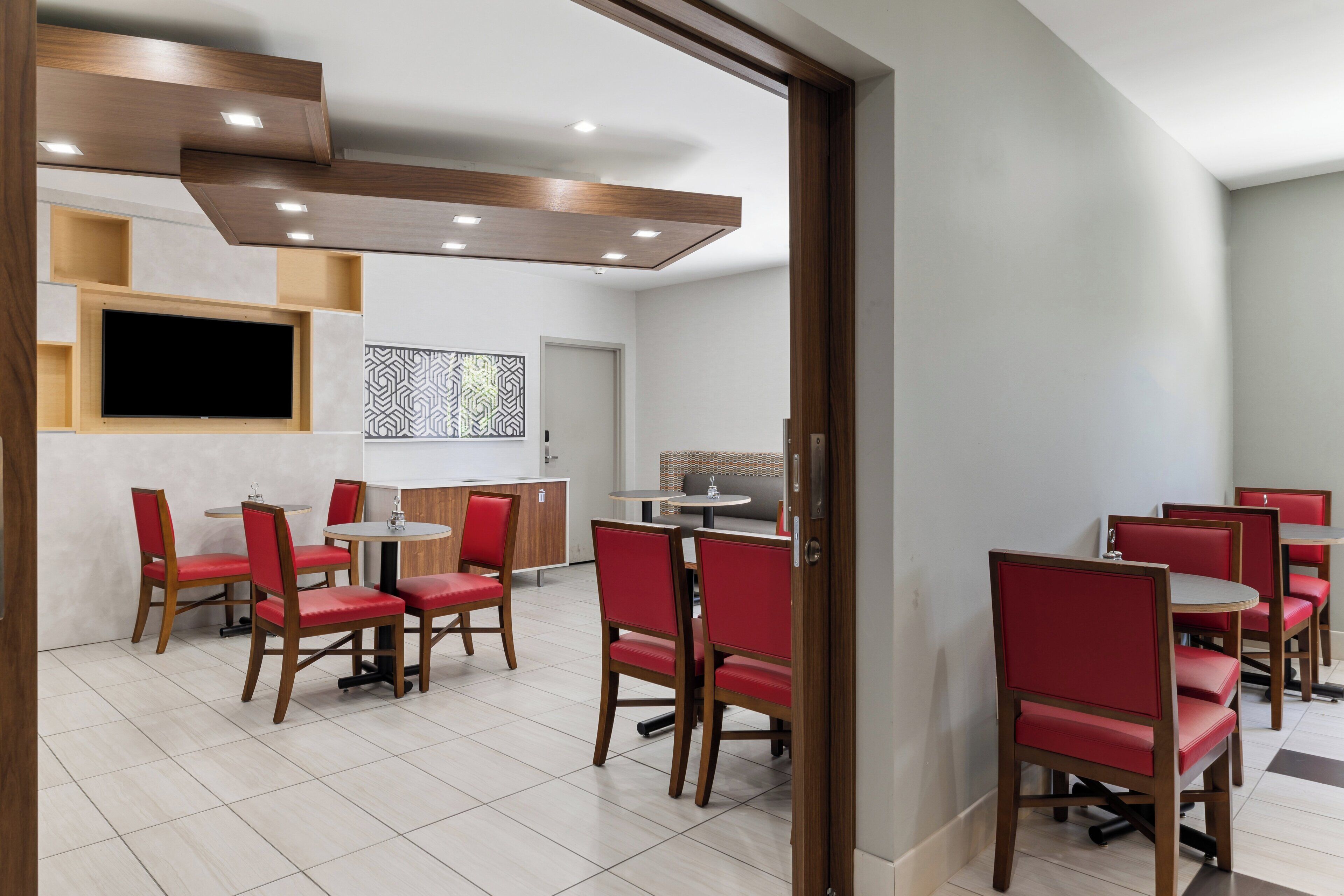 A modern dining area with red upholstered chairs, a wooden ceiling, and a flat-screen TV mounted on the wall.