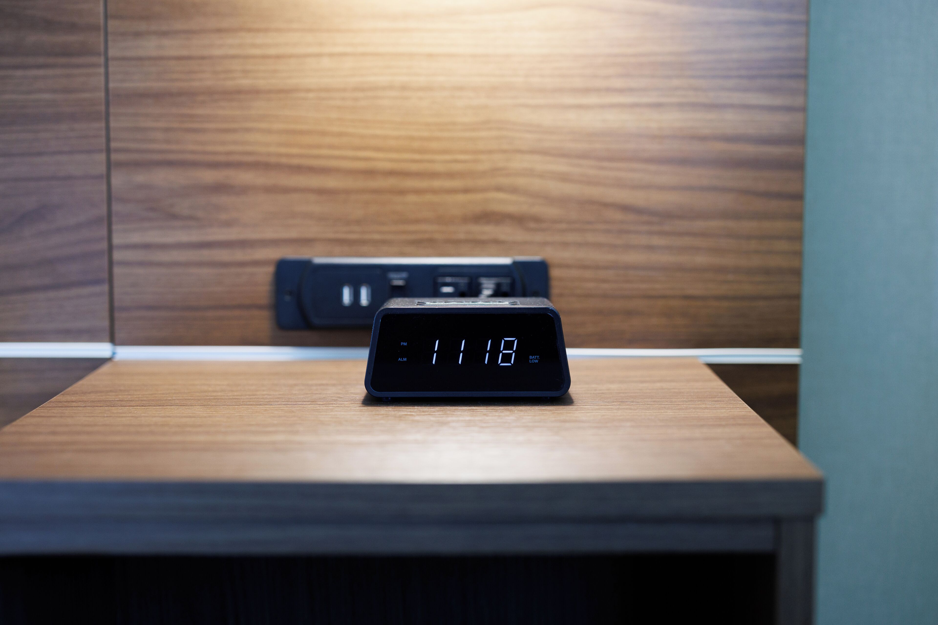 A digital clock on a wooden table in a room with a wooden panel wall.
