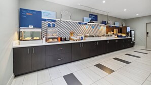 Una cocina moderna de hotel con una barra de desayuno, secciones identificadas para panadería, comida caliente y café, y un cartel que explica el servicio de desayuno.