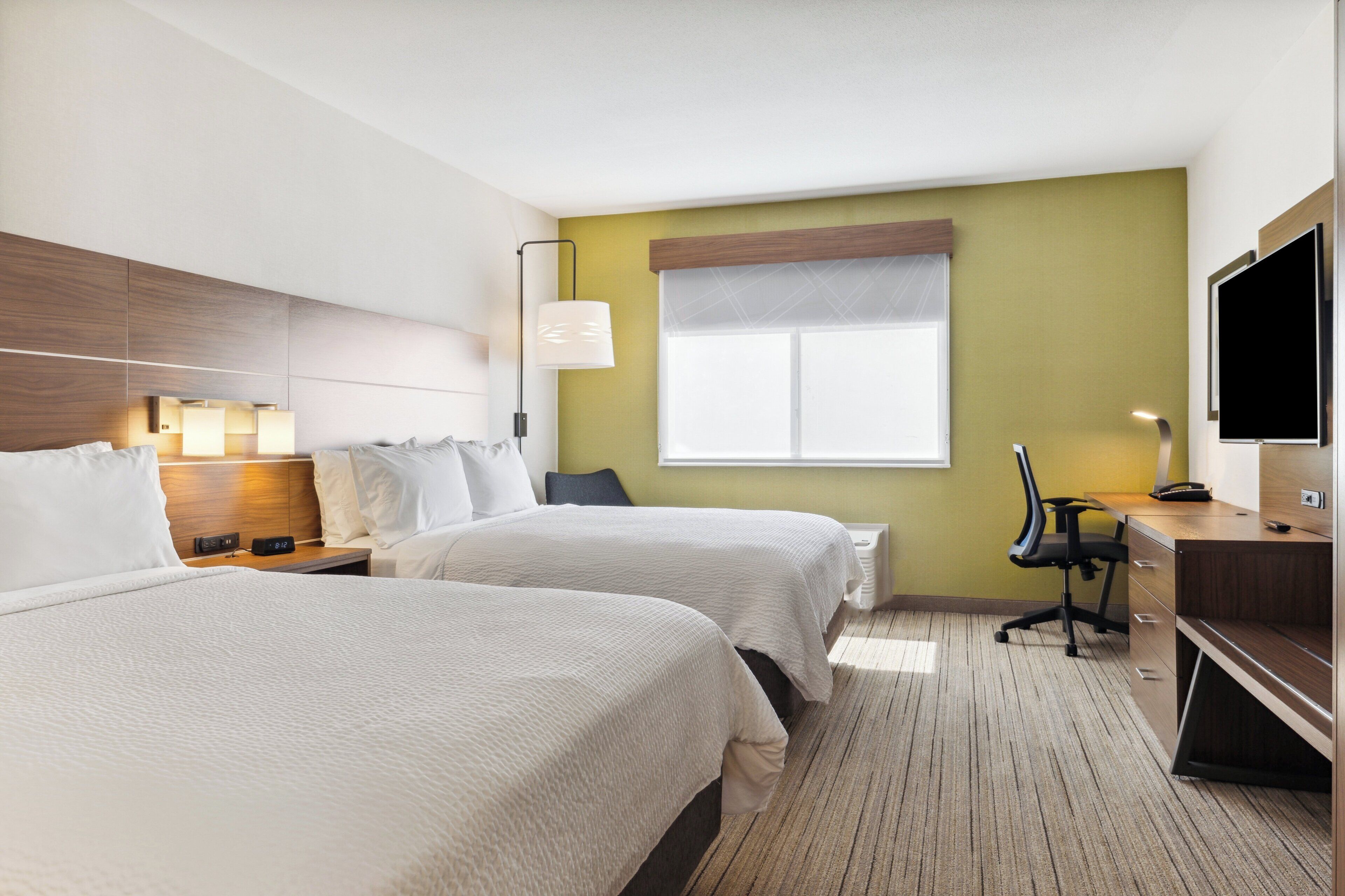A hotel room with two beds, a desk, a chair, a TV, and a window.