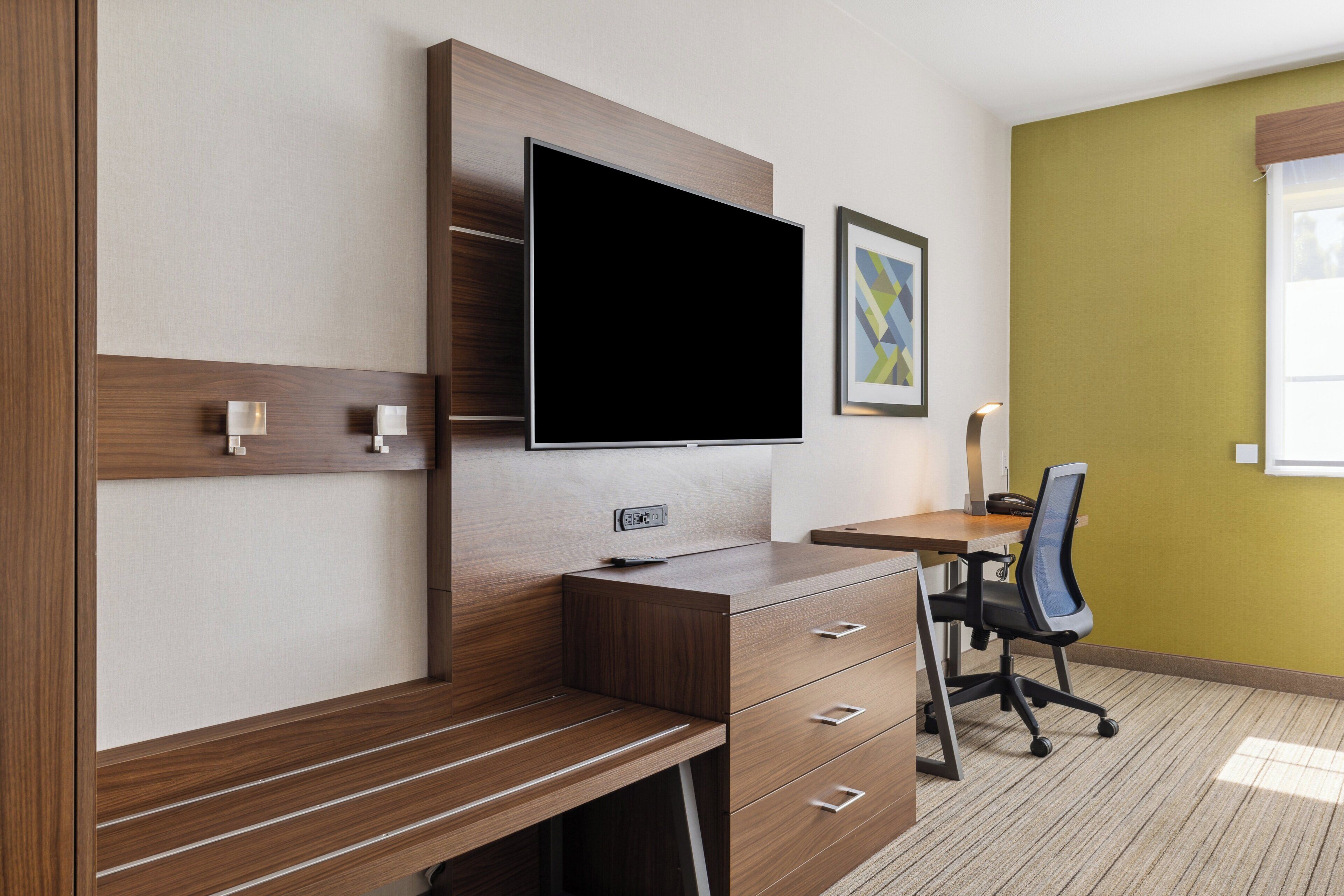 A modern hotel room with a wooden desk, a flat-screen TV, a chair, and a framed picture on the wall.