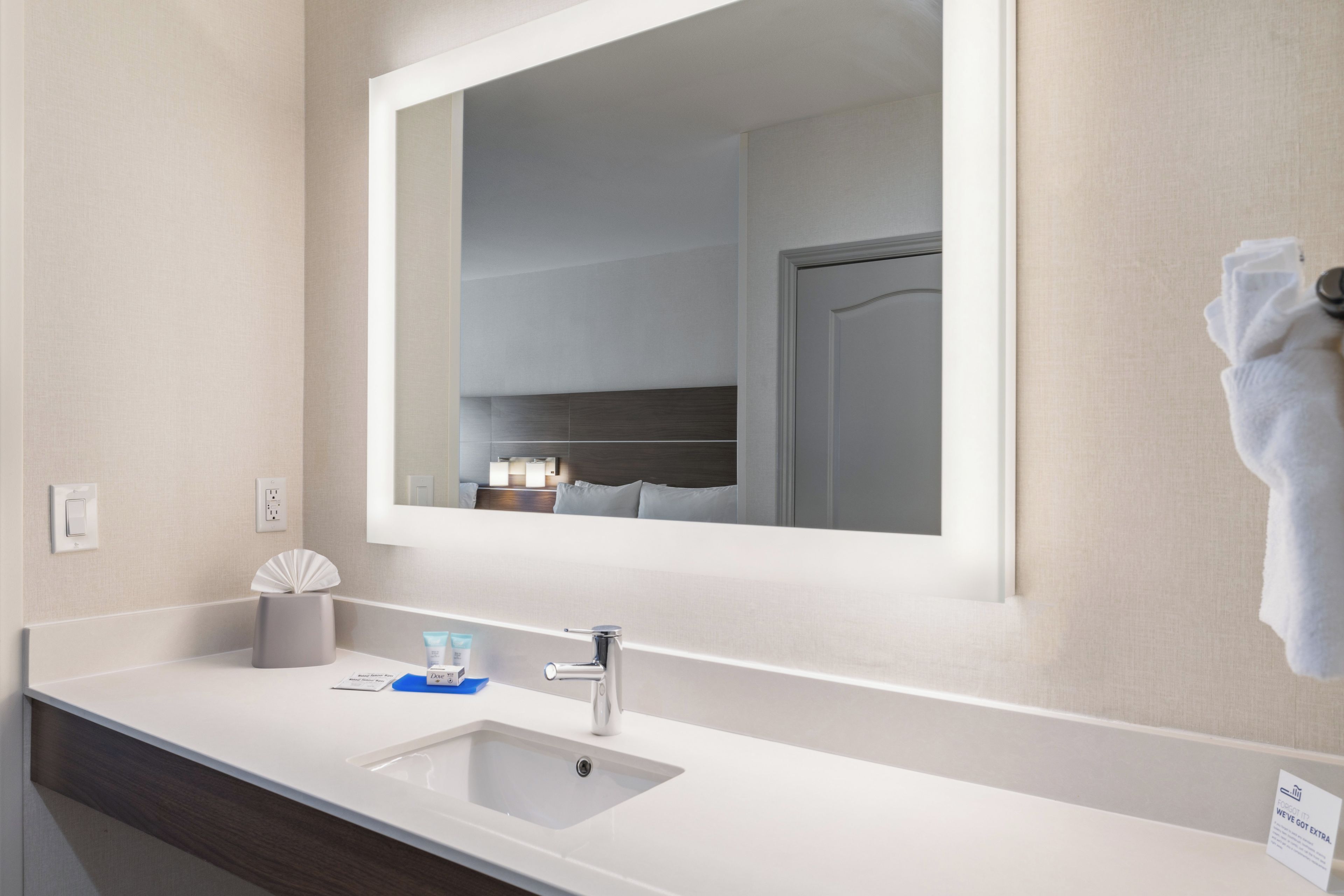 Une salle de bain d’hôtel avec un grand miroir, un lavabo, un porte-serviettes et un distributeur de mouchoirs.