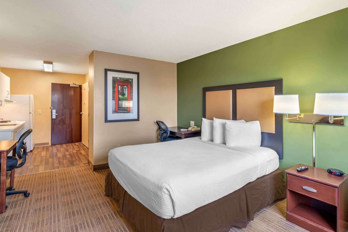 A hotel room with a large bed, a desk with a chair, a small table, and a nightstand with lamps.