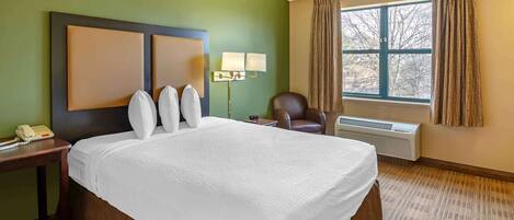 A hotel room with a large bed, a desk with a telephone, a chair, a window with curtains, and a wall-mounted lamp.