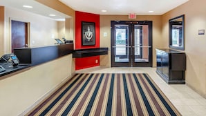 Et hotel lobby med en receptionsdisk, en rød accentvæg, et indrammet kunstværk, et stribet tæppe og en dør til udendørs.