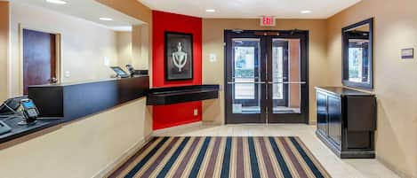 Vestíbulo de hotel con mostrador de recepción, pared decorativa roja, obra de arte enmarcada, alfombra a rayas y puerta doble que da al exterior.
