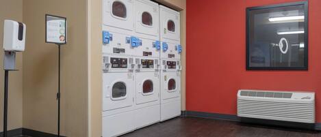 A laundry area with multiple washing machines and dryers, a hand sanitizer dispenser, and a red wall.