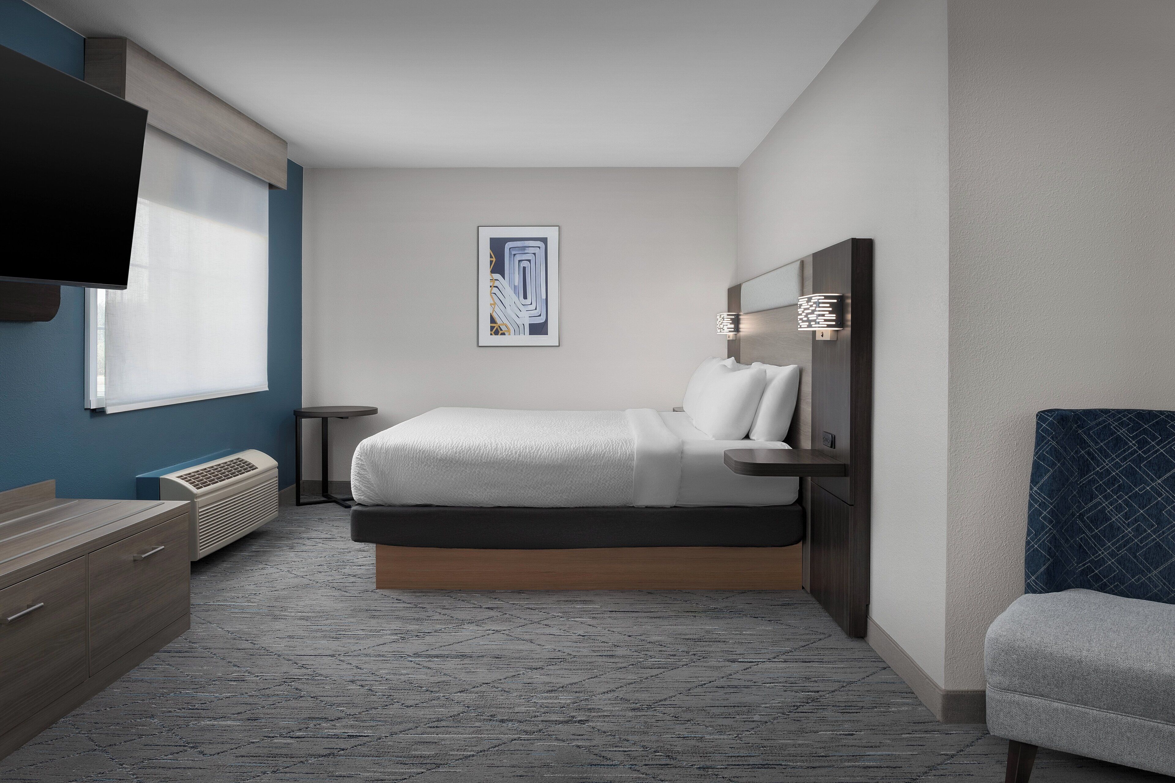A modern hotel room with a bed, bedside tables, a flat-screen TV, a wall-mounted artwork, and a window with blinds.