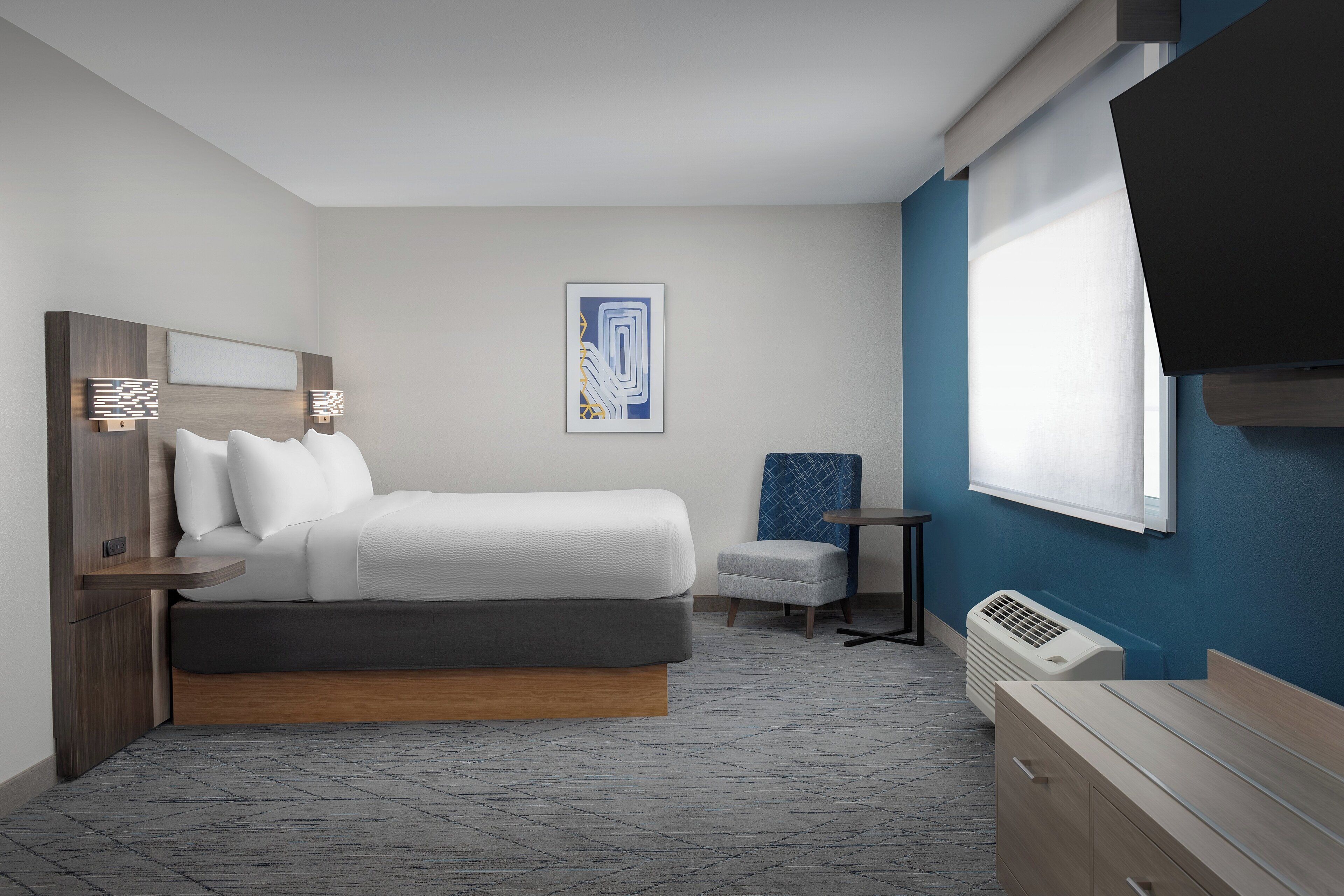 A modern hotel room with a bed, a small seating area, a TV, and a window.