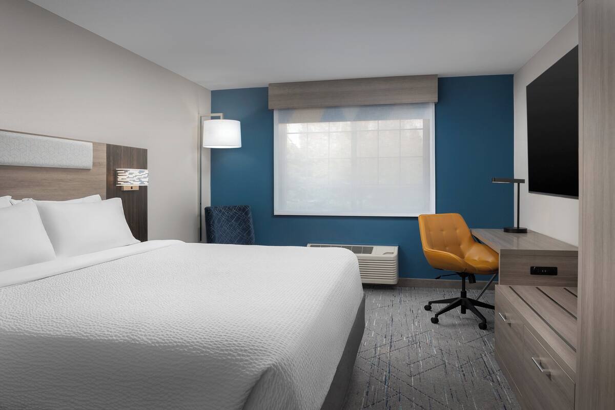 A hotel room with a large bed, a desk with a chair, a window with blinds, and a wall-mounted TV.