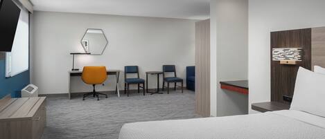 Quarto de hotel com cama, escrivaninha com duas cadeiras, mesa pequena e televisão.