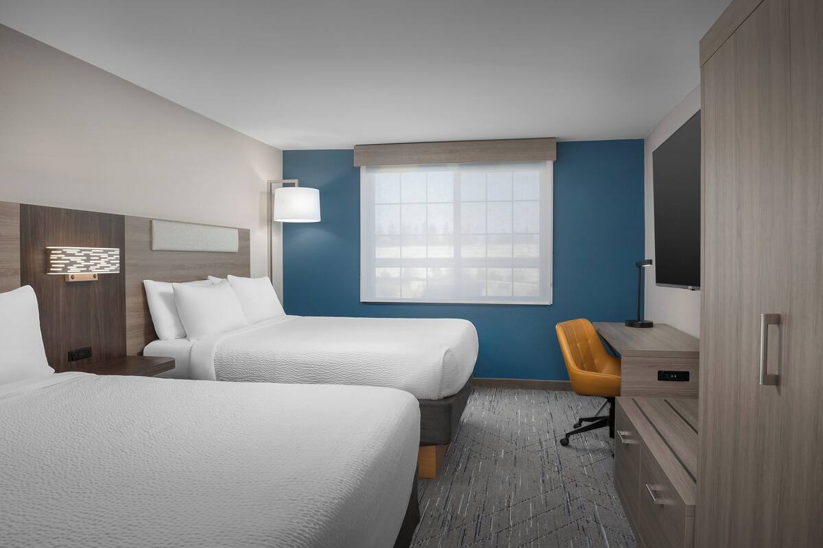 A hotel room with two beds, a desk, a chair, a TV, and a window with blinds.