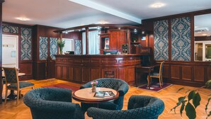 A hotel lobby with a curved reception desk, patterned wallpaper, and a wooden floor. There are several upholstered chairs and a small table with a vase of flowers. - Hotel Elysee (Prague)