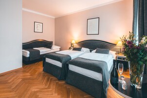 Habitación de hotel con dos camas, suelo de madera en diseño de espiga, mesita de noche con lámpara, florero con flores y cuadros en las paredes.