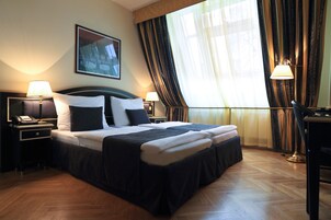 Quarto de hotel com uma cama grande, duas mesas de cabeceira com abajures, uma escrivaninha e uma janela com cortinas.