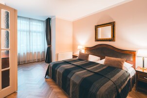 Quarto de hotel com uma cama grande, cabeceira de madeira, mesa de cabeceira com abajur, espelho na parede e vista para o corredor.