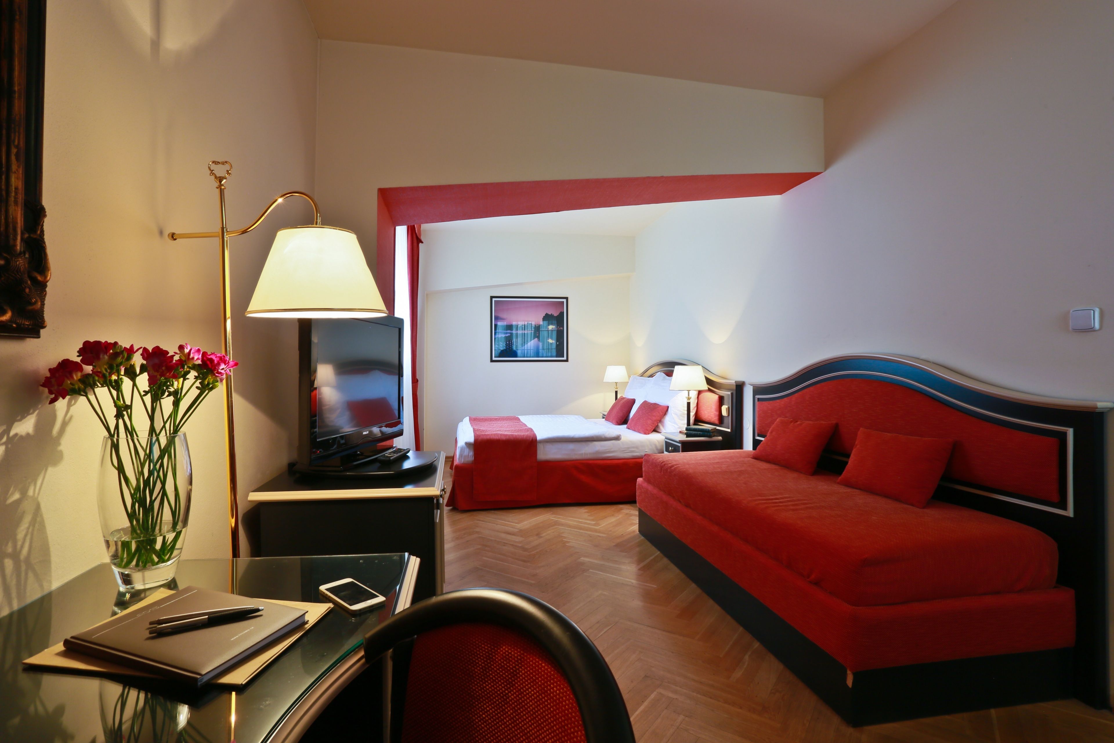 Habitación de hotel con una cama roja, un escritorio con lámpara, una silla, un jarrón con flores y un televisor.
