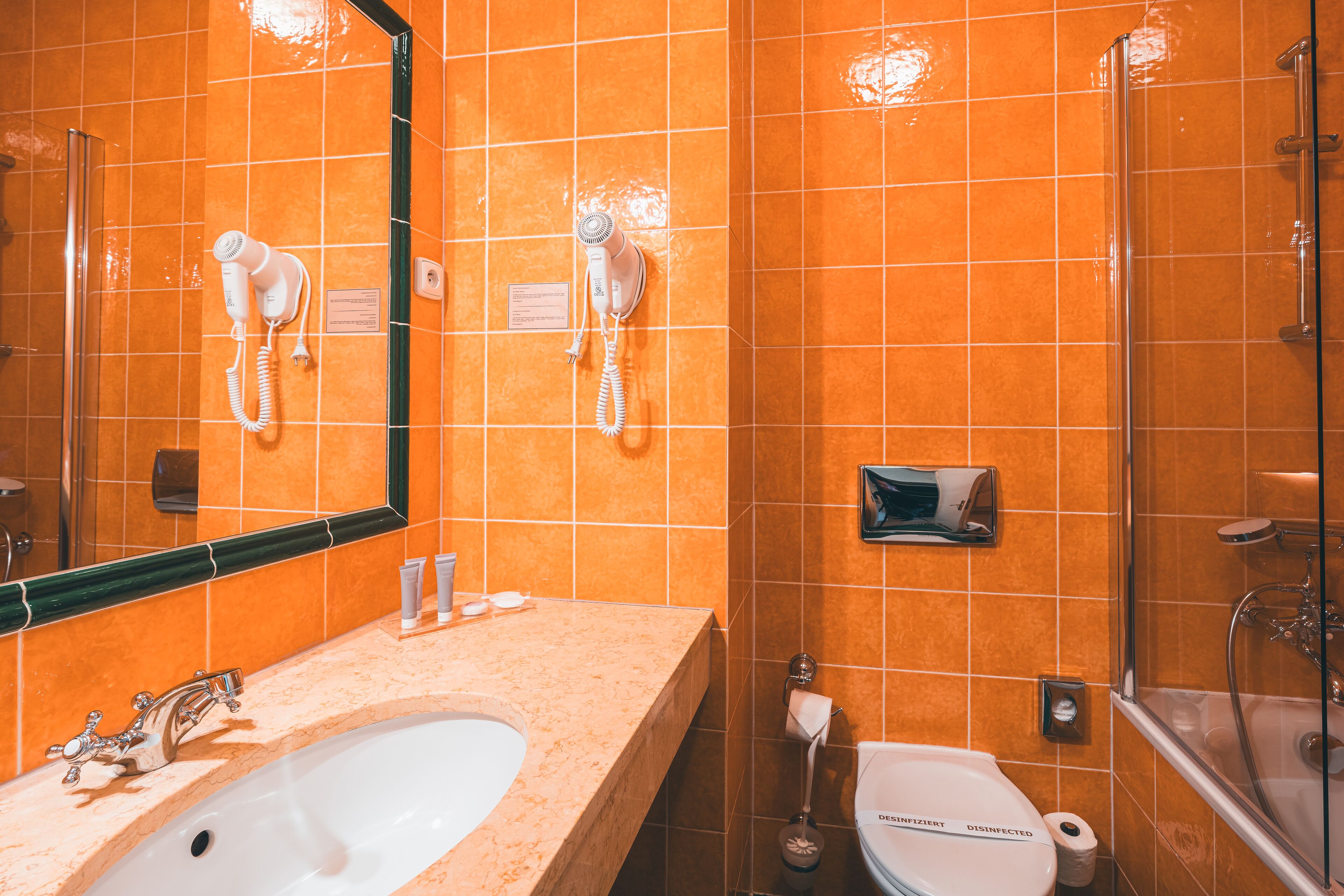 Casa de banho com azulejos laranja, uma sanita, um lavatório branco, um espelho, secador de cabelo e chuveiro.