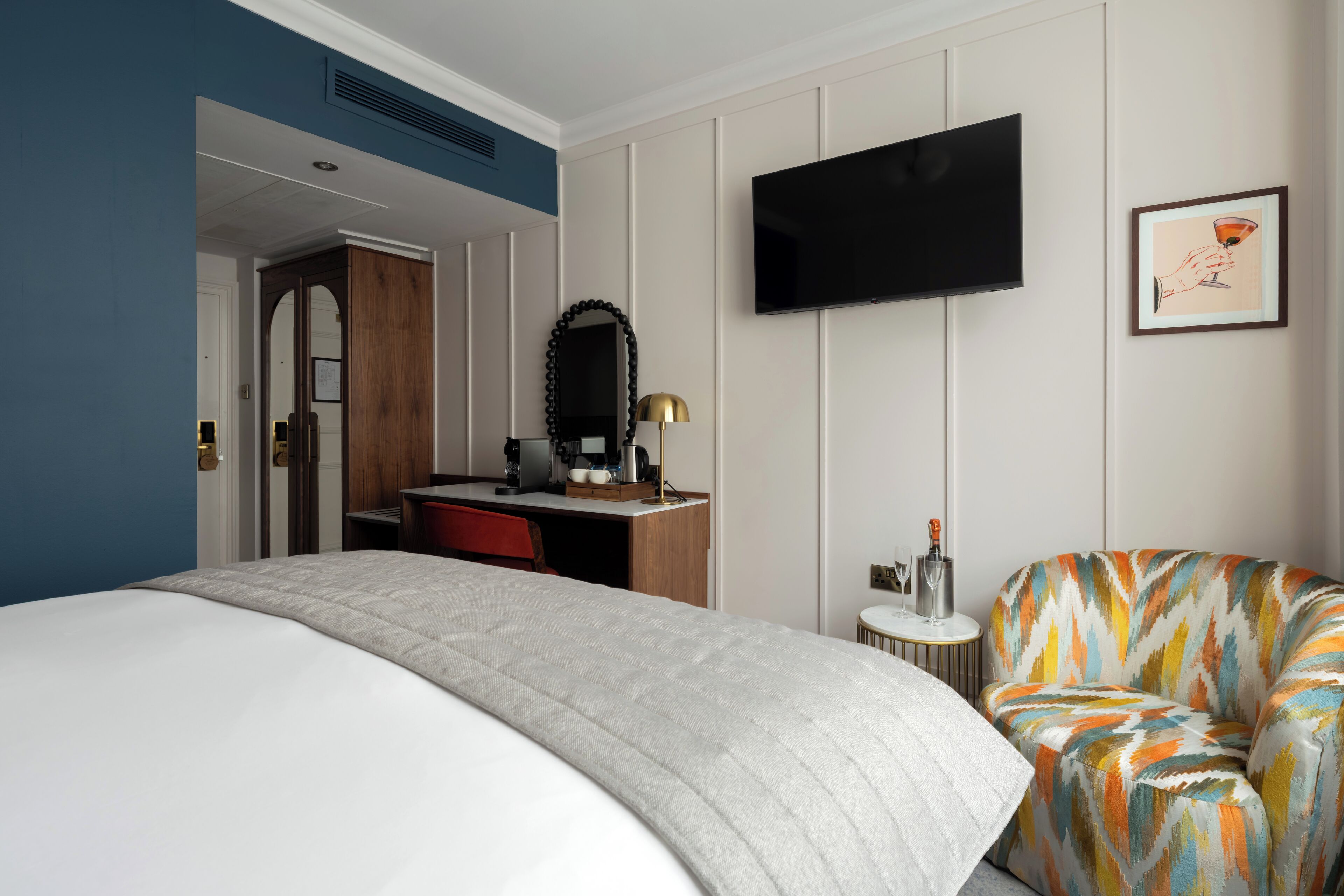 Une chambre d’hôtel avec un lit, un bureau doté d’un miroir, une télévision, un tableau encadré et un fauteuil coloré.