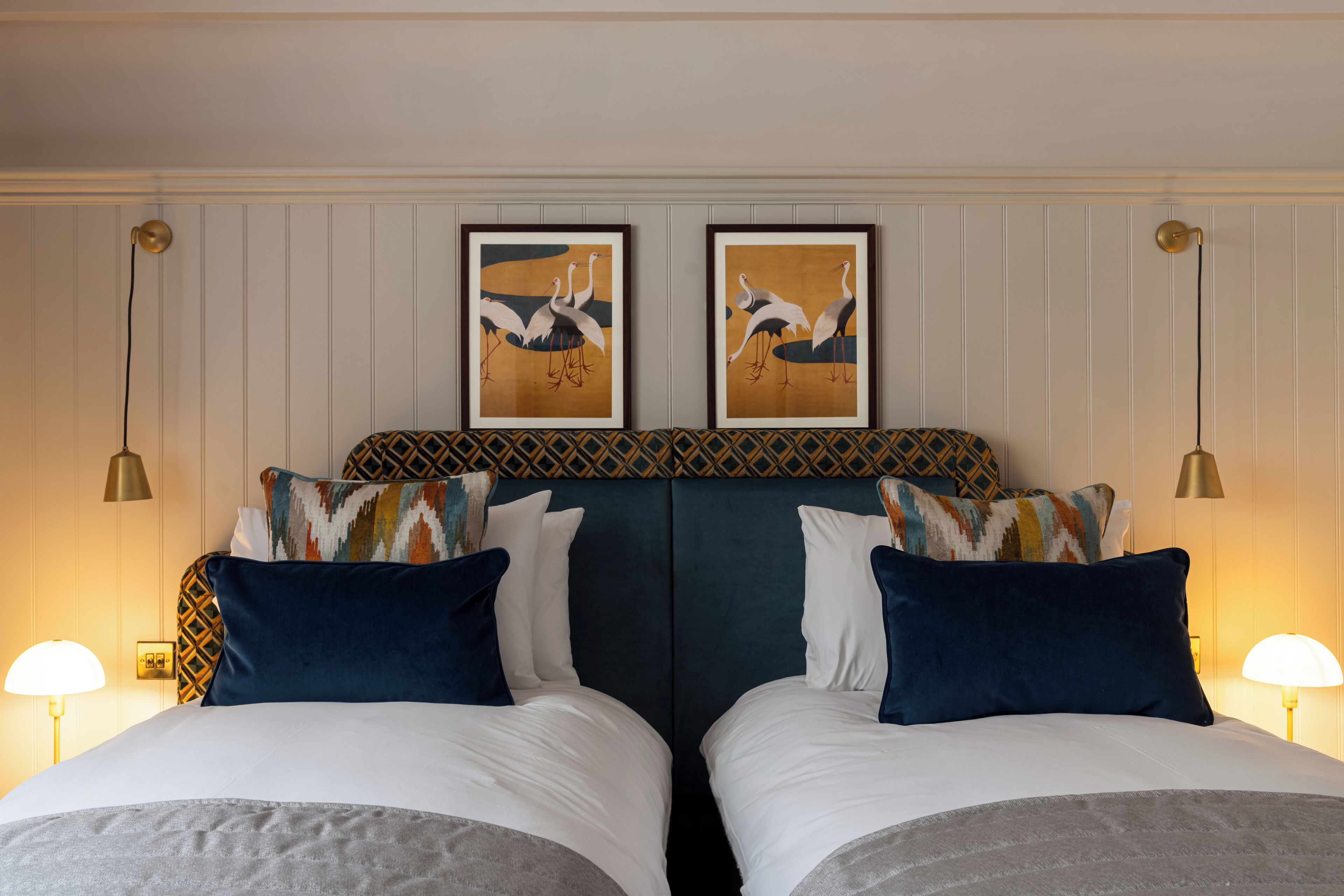 A hotel room with two beds, each with a patterned headboard, white bedding, and decorative pillows. There are wall-mounted lamps on either side of the bed.