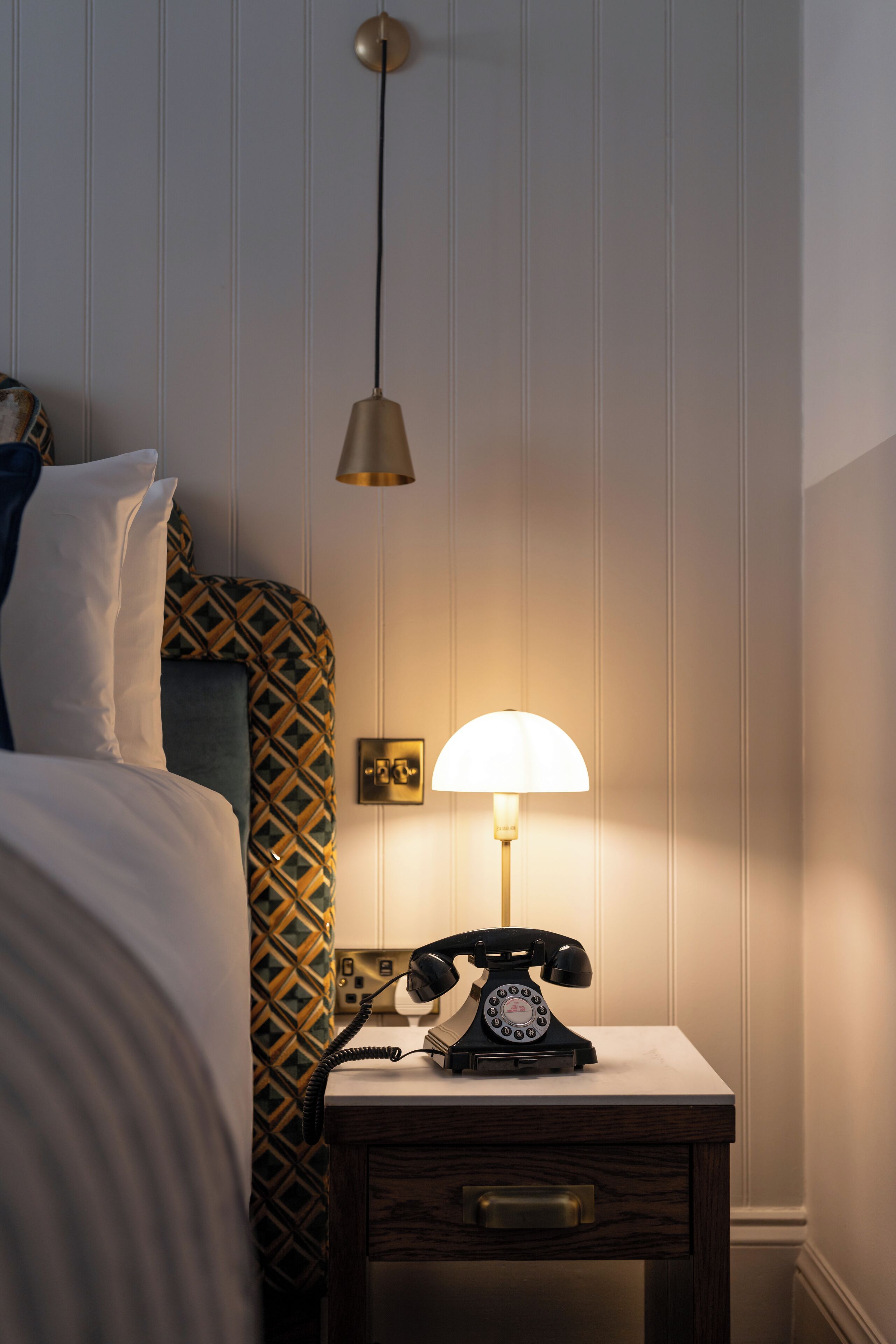 A hotel room with a bed, a nightstand with a telephone, and a wall-mounted lamp.