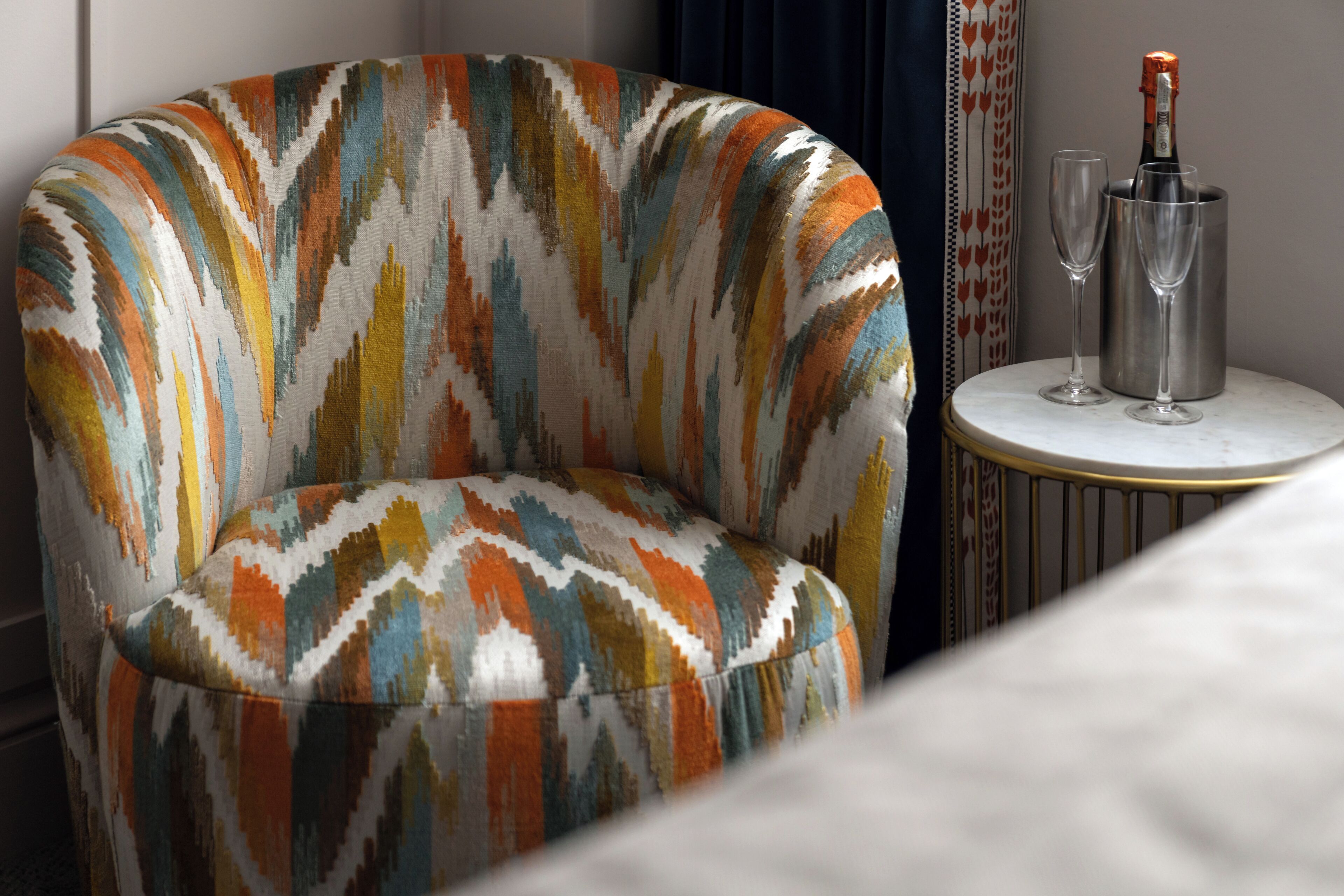 Un sillón moderno con un estampado geométrico y colorido, una mesita redonda con una botella de champán y dos copas, y una cortina oscura al fondo.