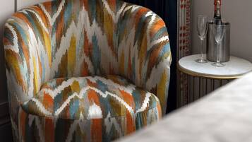 Un sillón moderno con un estampado geométrico y colorido, una mesita redonda con una botella de champán y dos copas, y una cortina oscura al fondo.