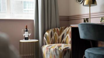 Une pièce comprenant un fauteuil à motifs, une petite table ronde avec une bouteille de vin et des verres, un lampadaire et une fenêtre avec des rideaux.