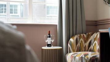 Una habitación con ventana, un sillón con estampado, una mesita redonda con una botella de vino y dos copas, y un espejo en la pared.
