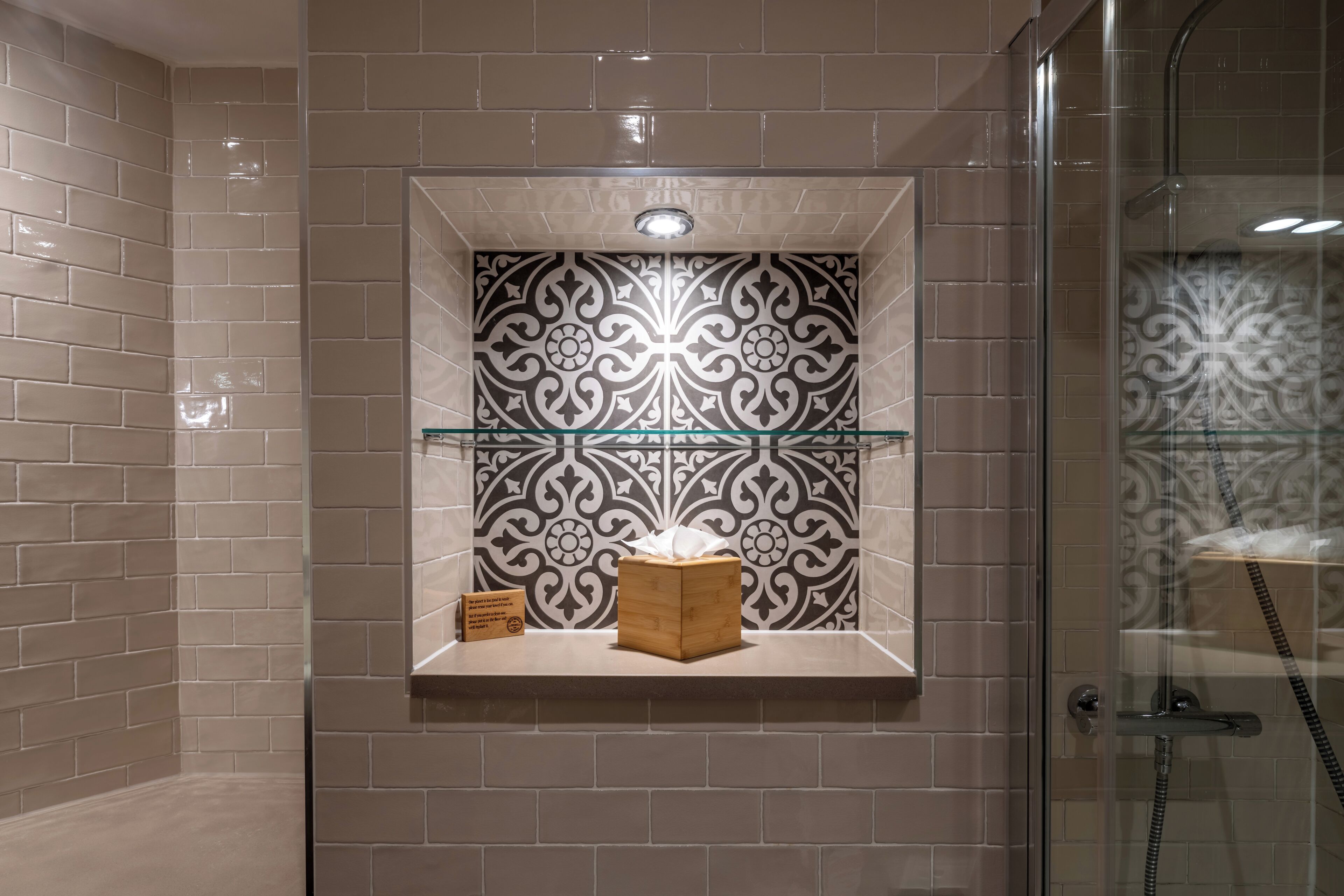 Un baño con baldosas color beige, una pared decorativa con azulejos blanco y negro, un caja de madera y una estantería de vidrio.